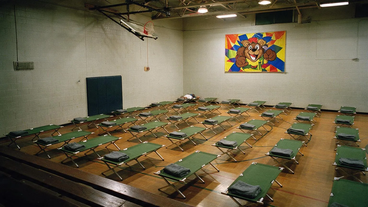A school gymnasium filled with rows of emergency cots beneath a cheerful laminated sports banner.